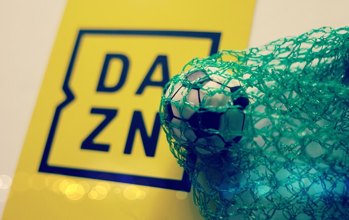 Ein Fußball liegt im Netz auf einem DAZN-Logo