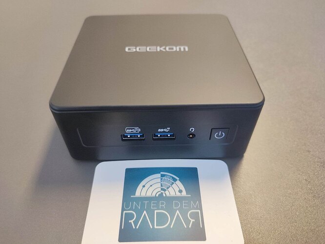 Der GEEKOM IT15 steht auf einem Tisch. Im Vordergrund eine Visitenkarte von Tarnkappe.info.
