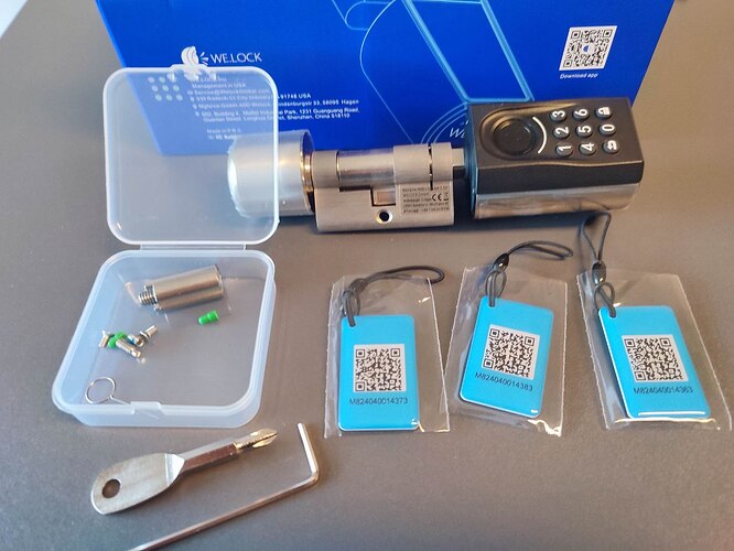 Nahaufnahme des vollständigen Lieferumfangs des WELOCK Touch A51 Smart Locks: Auf dem Bild sind das Türschloss mit Tastenfeld, eine geöffnete Kunststoffbox mit Montagematerial, drei RFID-Karten, ein Innensechskantschlüssel und ein kleiner Schraubendreher vor der Produktverpackung zu sehen.