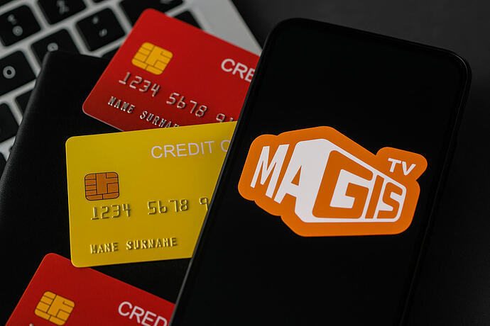 Auf einem Laptop liegen ein Smartphone mit MagisTV-Logo und rote sowie gelbe Kreditkarten. Das Bild symbolisiert die Gefahr des Datenklaus durch Malware in Streaming-Apps.
