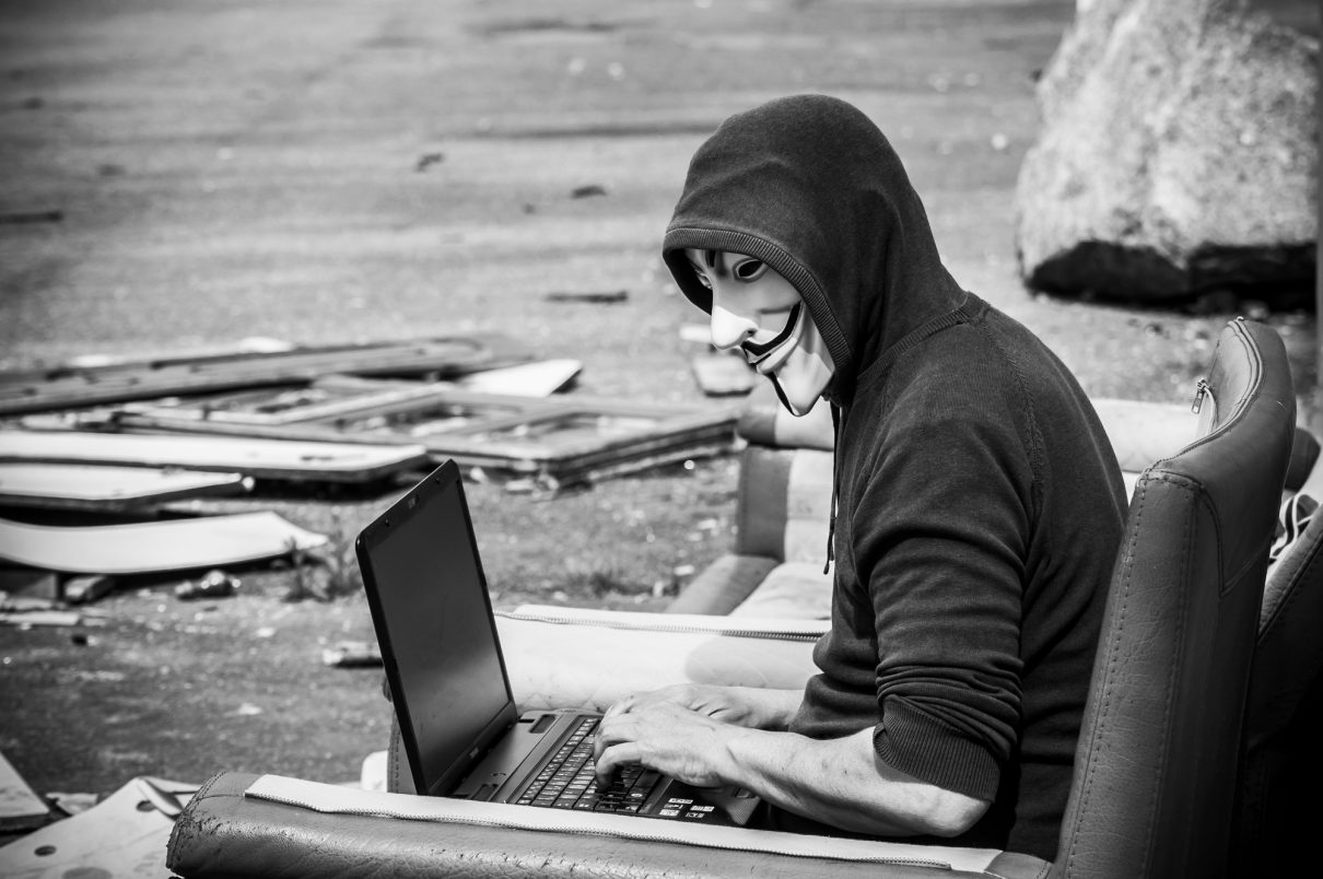 Porträt eines Hackers mit Vendetta-Maske und Computer in einer verlassenen Fabrik auf einem alten Sessel