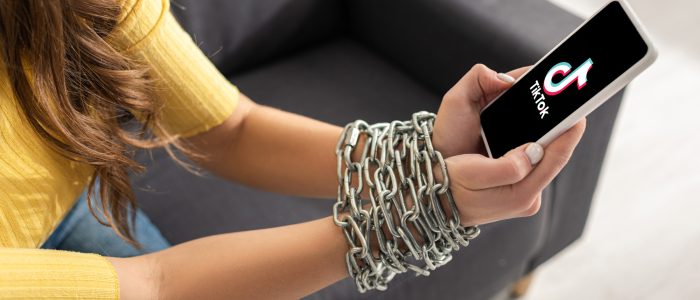 Eine junge Frau, die auf einem Sofa sitzt und ihr Smartphone mit der TikTok-App in den mit einer Metallkette gefesselten Händen hält