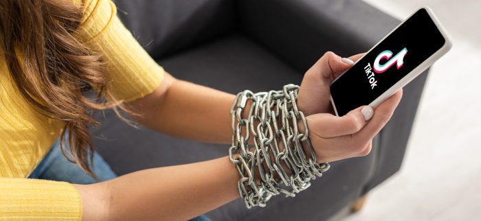 Eine junge Frau, die auf einem Sofa sitzt und ihr Smartphone mit der TikTok-App in den mit einer Metallkette gefesselten Händen hält