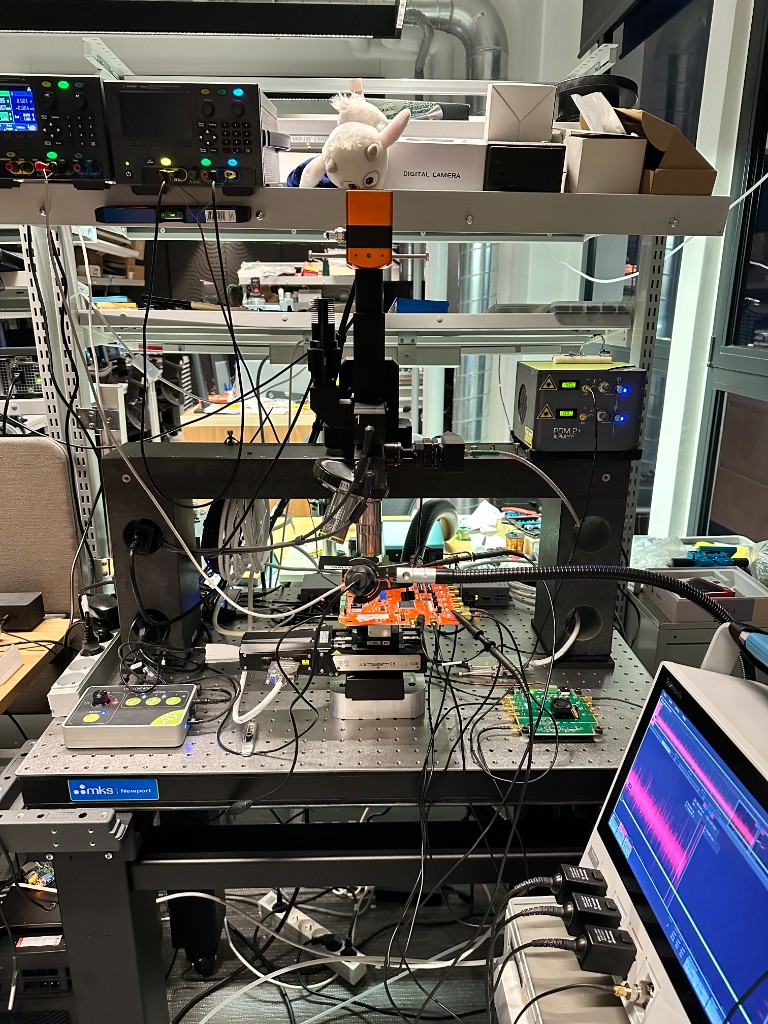 Laser‑Fault‑Injection‑Versuchsaufbau im Hardware‑Sicherheitslabor: optischer Tisch mit Mikromanipulator, Laseroptik und verdrahtetem Testboard; Oszilloskop und Netzteile zeigen Messsignale im Hintergrund; zahlreiche Messleitungen und ESD‑sicheres Setup, keine Personen sichtbar.