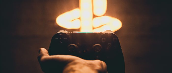 Das Bild zeigt einen ausgestreckten Arm mit einem Controller. An der Wand leuchtet das Symbol für Playstation von Sony.