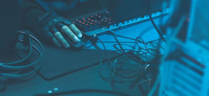 Ein unbekannter männlicher Krimineller mit fingerlosen Handschuhen sitzt an einem Schreibtisch mit Drähten und Kabeln und plant einen Ransomware-Angriff
