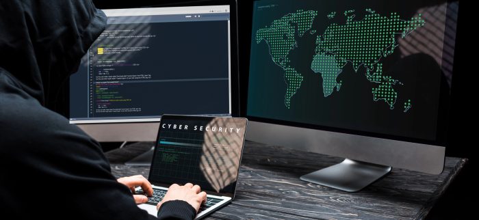 Hacker mit Laptop und Cybersecurity-Schriftzug auf dem Bildschirm neben Computermonitoren