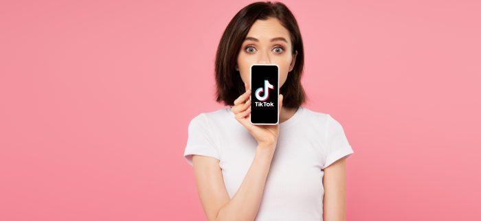 Mädchen mit überraschtem Blick hält sich ein Smartphone mit TikTok-Logo vor den Mund